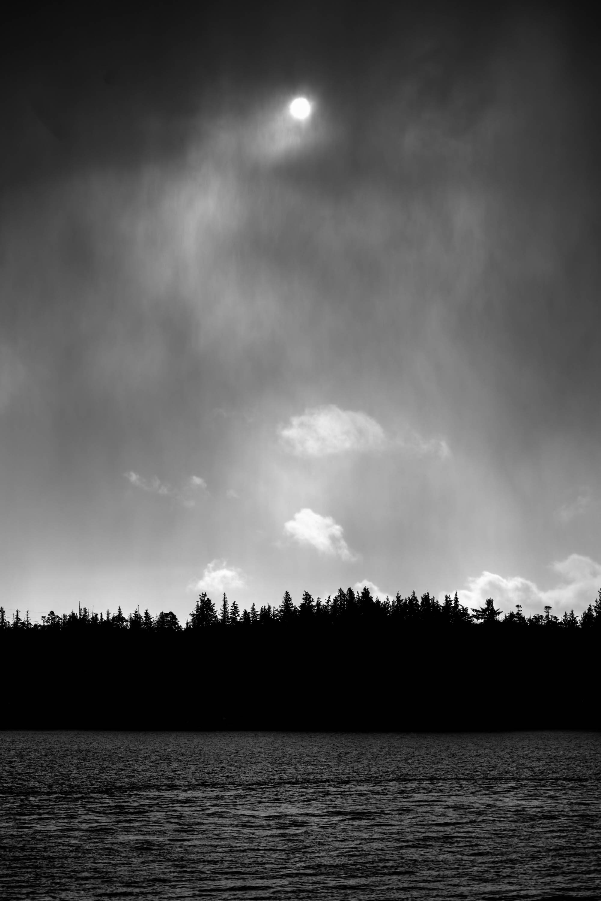 Between the viewer and the sun is an island covered in darkness. A raincloud pouring down on the island creates a gradient of grey above the island while allowing bright clouds to be seen behind the rain.