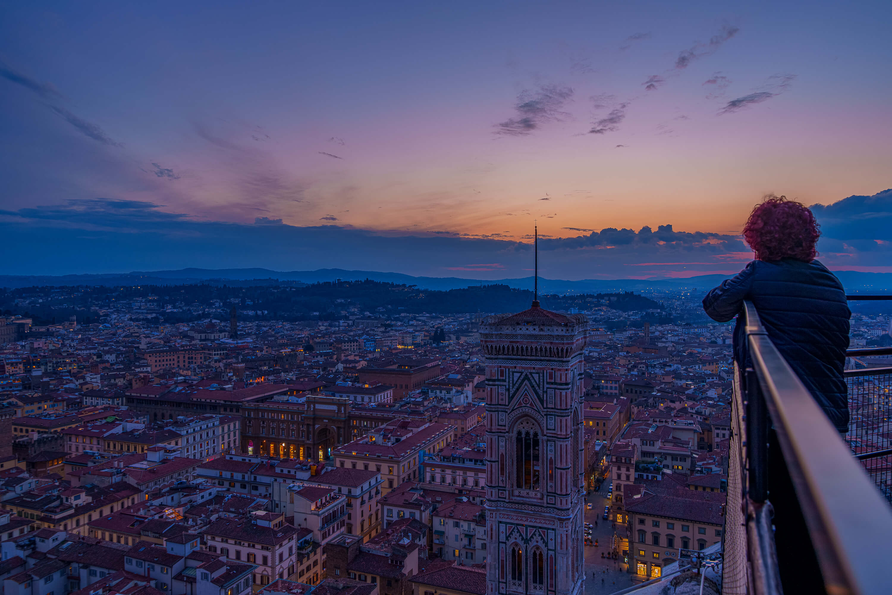 Poised atop the Duomo in Firenze, a visitor looks out at a city bathed in blue hour light. Light orange, purples, and blues cover the scene both from natural light and artificial city light.