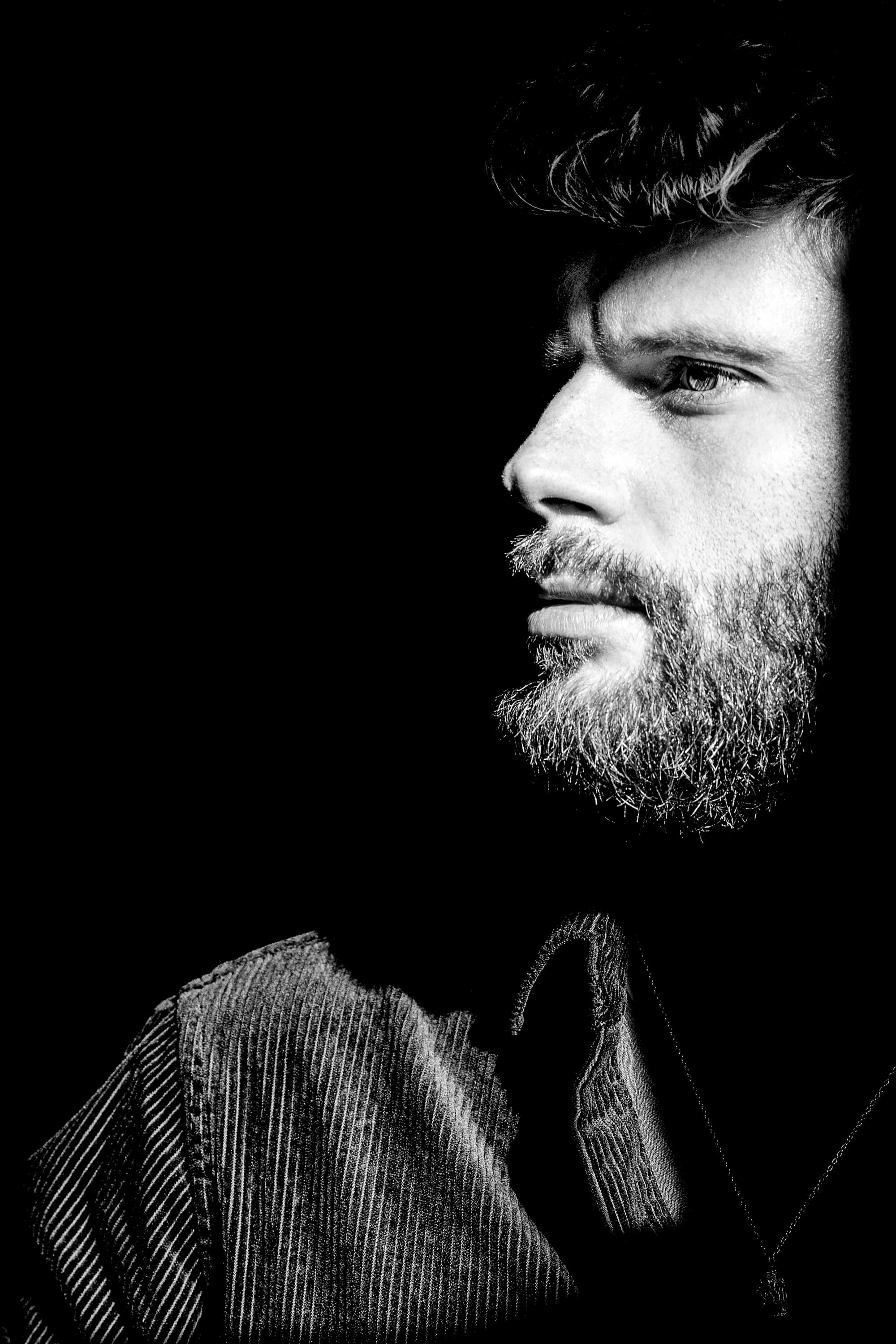 A model is bathed in angled light showing the texture on his face, shirt, and necklace. The rest of the photo is covered in darkness.