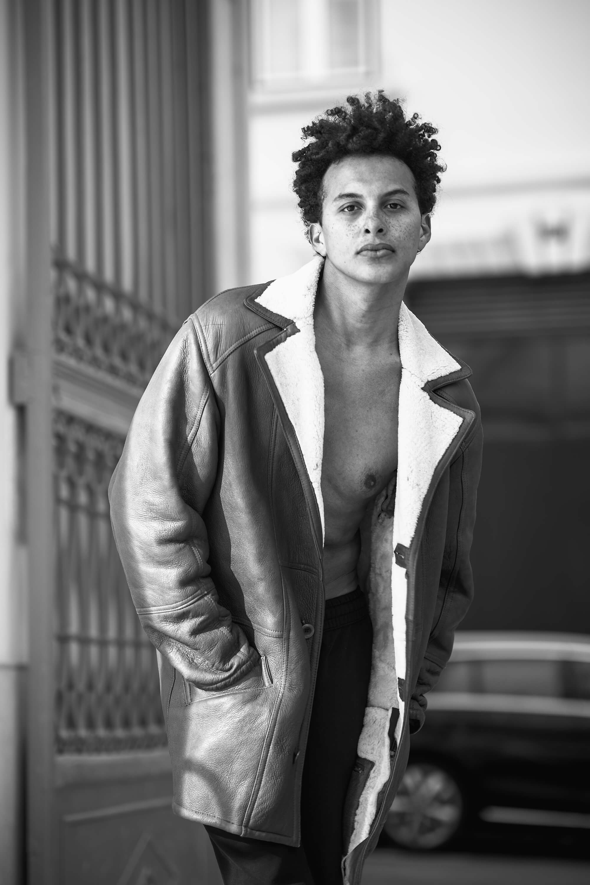 A model stands by an open gate. He poses shirtless wearing a jacket.