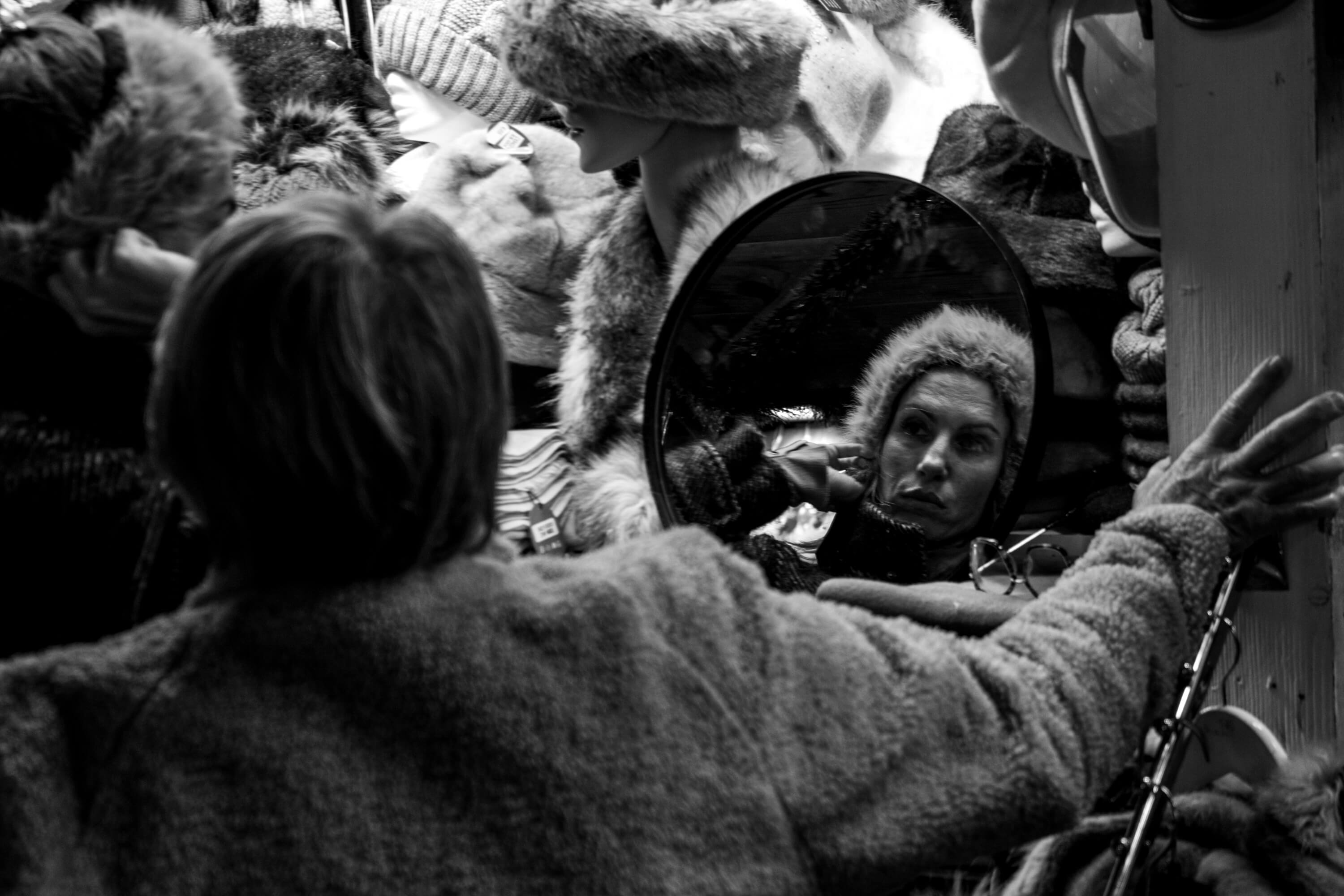 A woman tries a hat on at a market stall. We see her through the reflection of the mirror she is using to view herself.