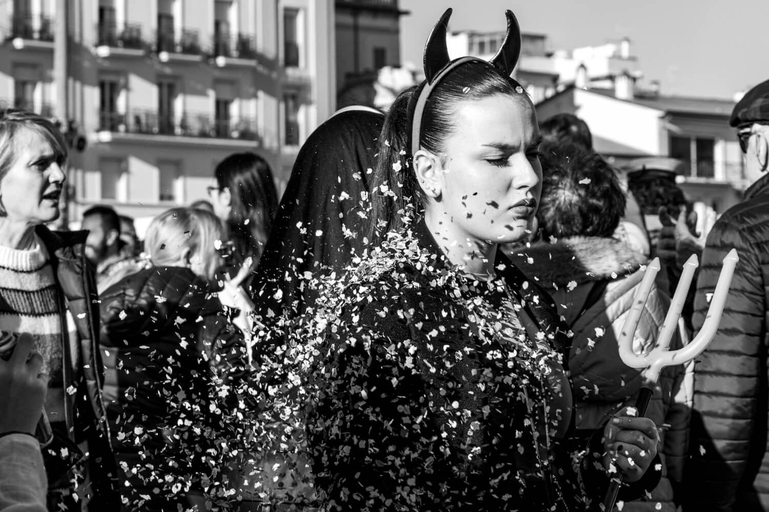 A woman in devil horns wielding a trident walks in Viareggio. She is surrounded by confetti but appears unfazed and disinterested.