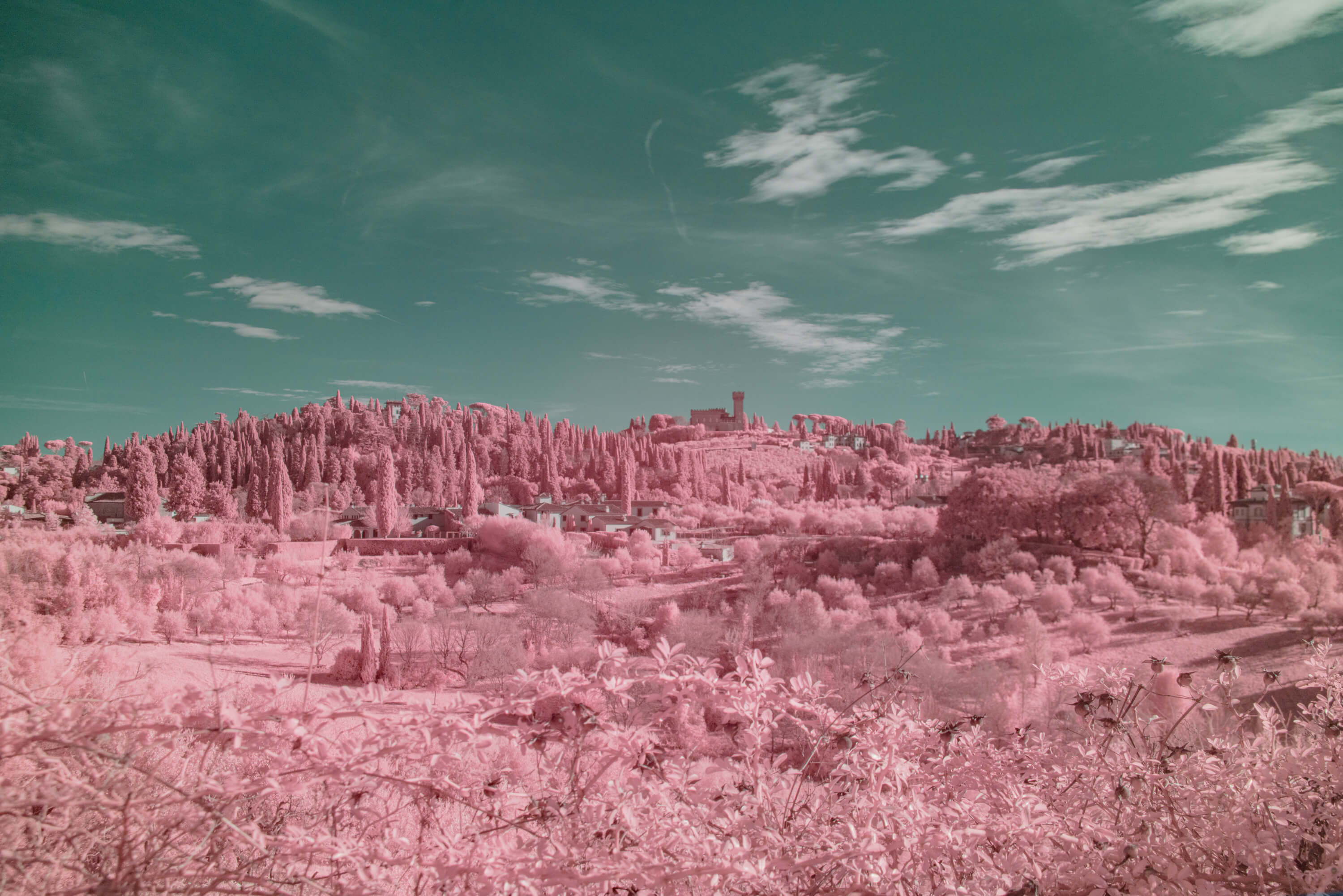 A Tuscan hillside studded with villas, towers, and cypress trees is shot in infrared. Foliage blooms pink across the slope while the sky turns seafoam green, making the stone buildings appear to float in a pastel landscape.