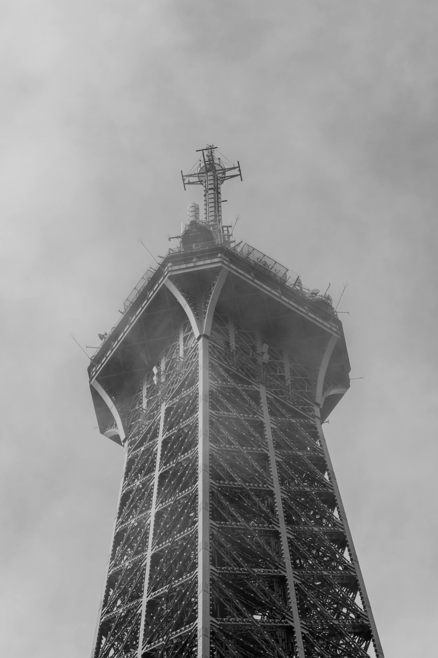 The tip of the Eiffel Tower is covered by a rolling mist. Slightly obscured, the various wires and communication devices can still be seen protruding in every direction.