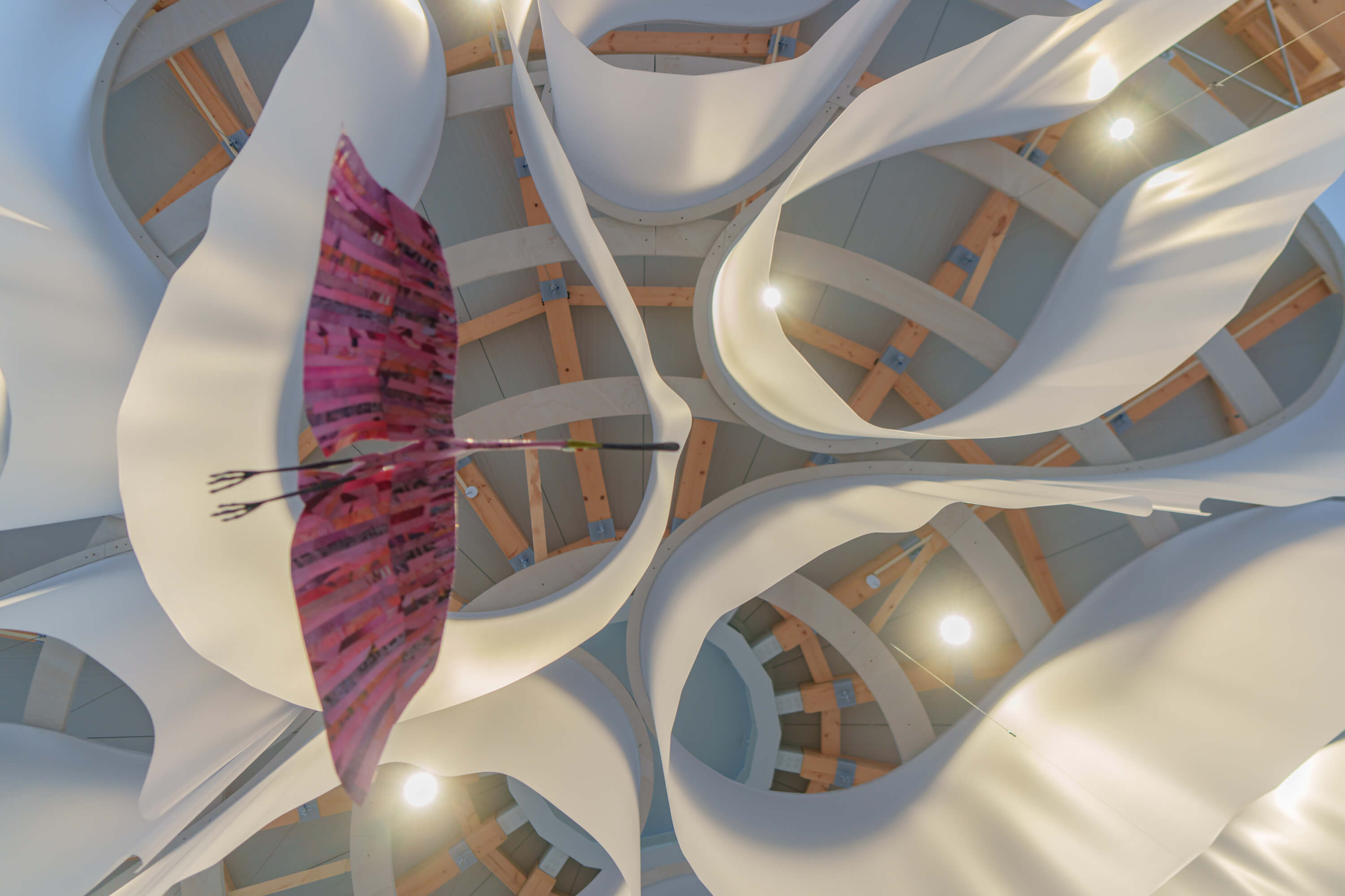 A paper flamingo is suspended above the viewer. Viewed from below, the paper that represents the clouds becomes a winding pattern and the wooden structure the installation is attached to can be seen.