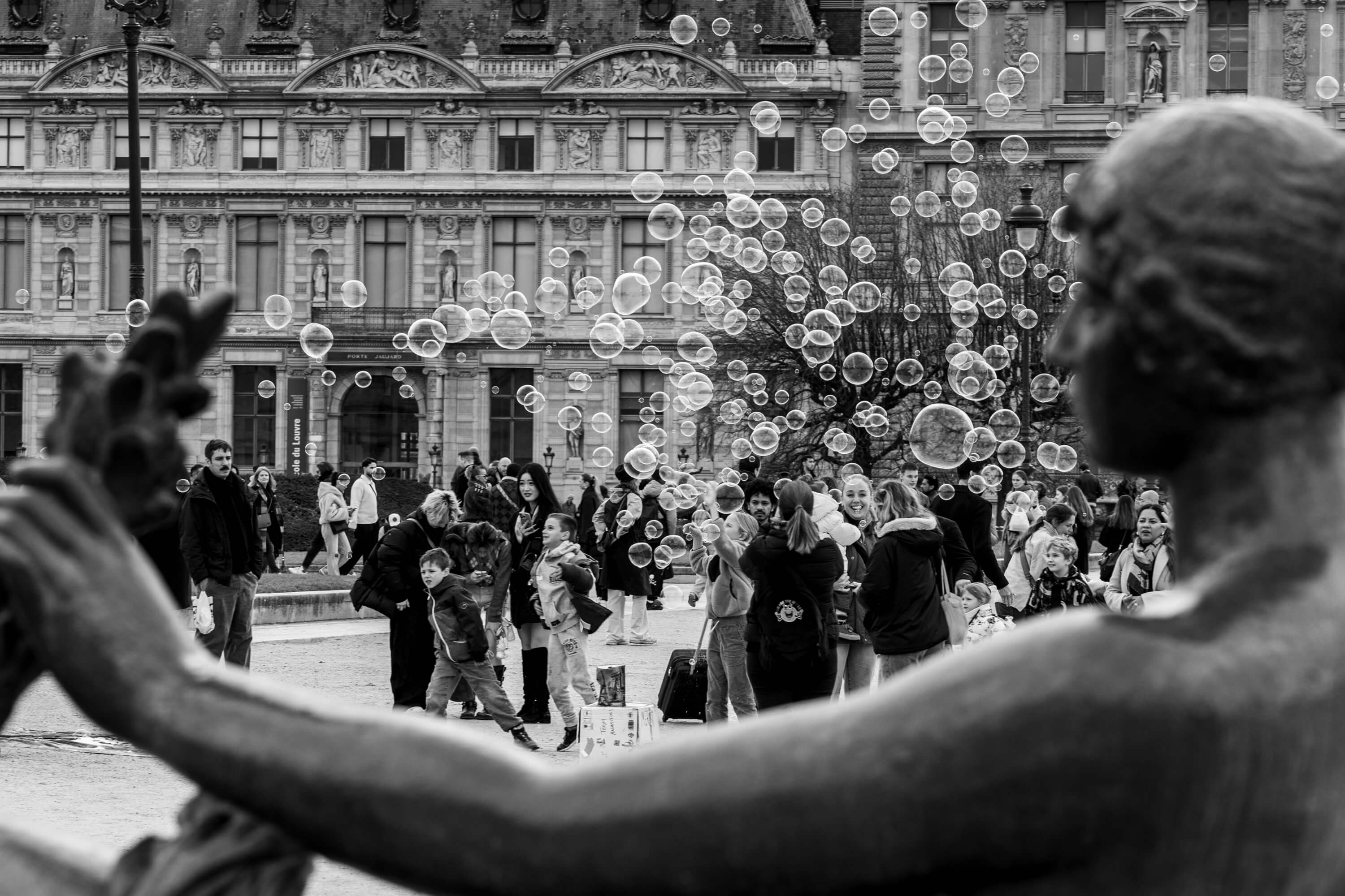 A statue frames the scene where bubbles float around a crowd in the Jardin du Carrousel. A wing of the Louvre makes up the background.