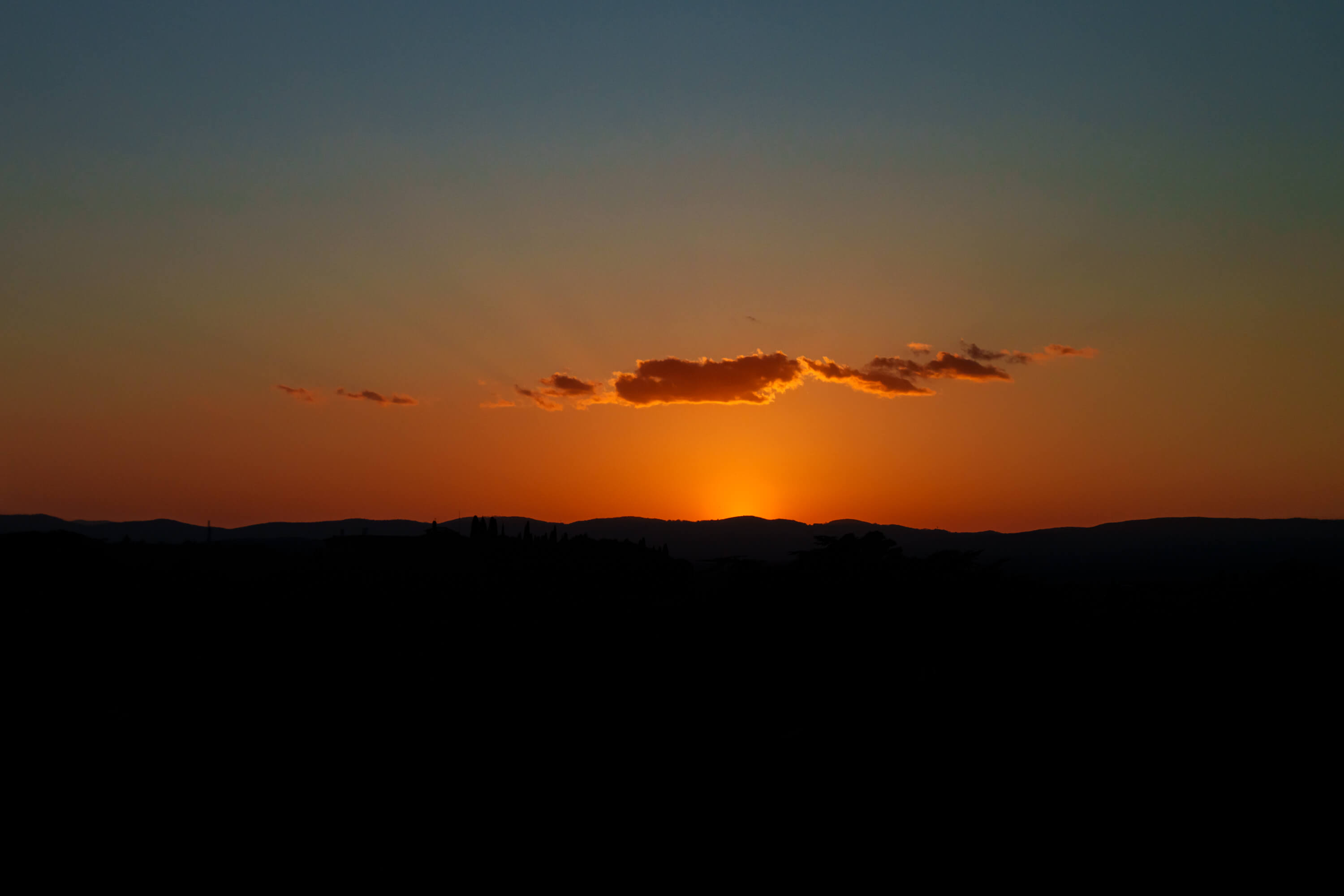 Golden light shines from below a line of hills upwards into a grouping of clouds. The hills are covered in shadow while the light transitions upwards from golden orange into a pale blue.