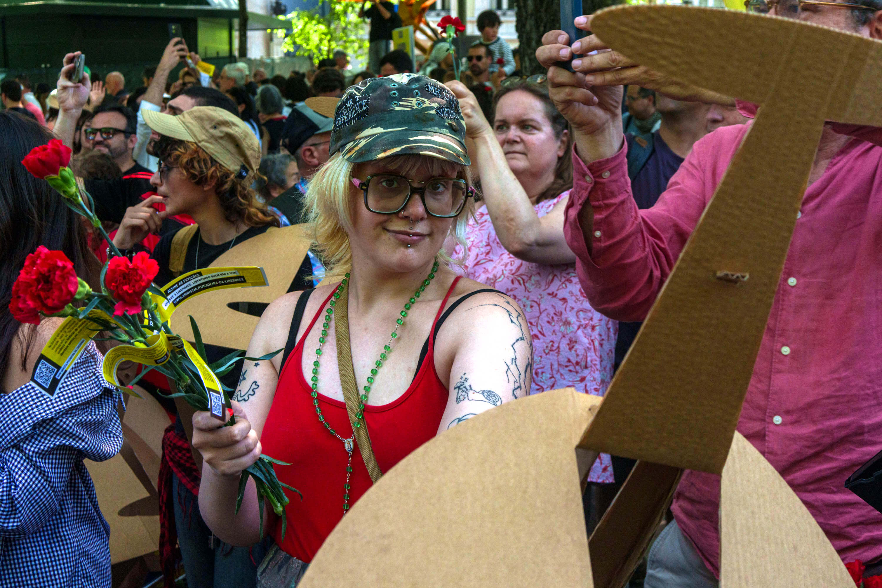 A woman in red attempts to hand the photographer a cardboard object. She is holding red carnations in celebration of the 52nd anniversary of the Carnation Revolution in Portugal.