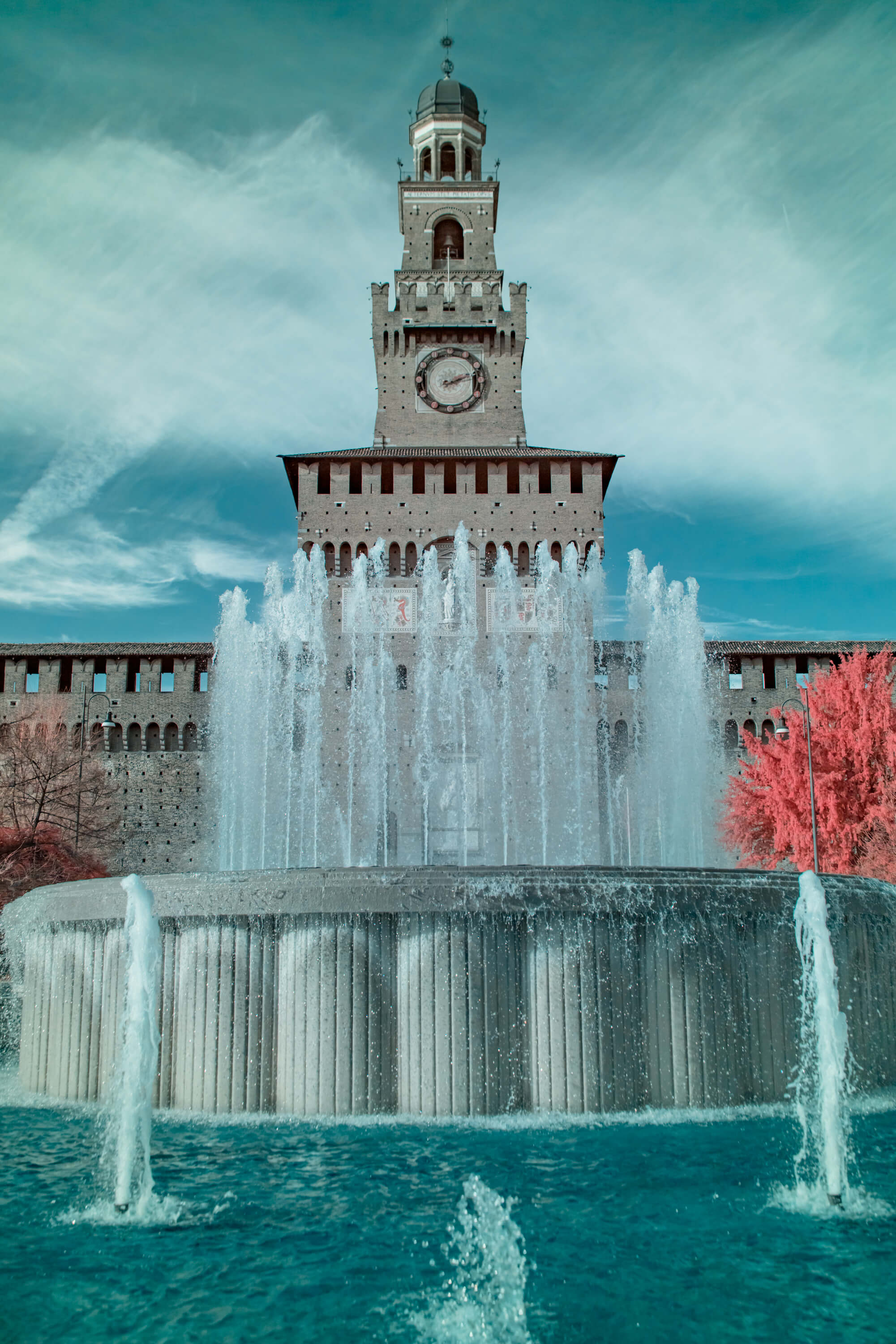 The fountain of Piazza Castello shoots turquoise water in front of the clock tower of Castello Sforzesco, Milan. Infrared turns the sky and water a vivid teal while a lone tree glows hot pink to the right.