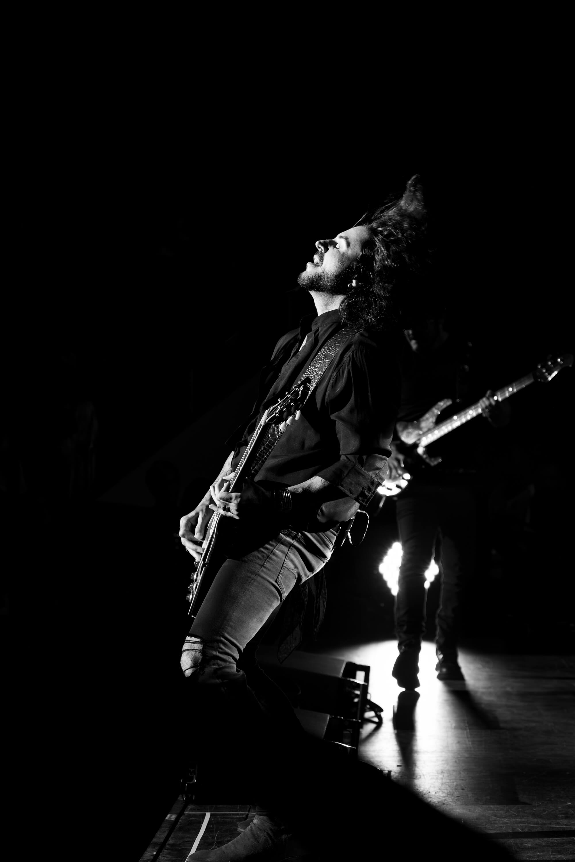 Marco Túlio of the Brazilian pop rock band Jota Quest appears lost in the moment. Hair flung back while a spotlight shines directly on him.