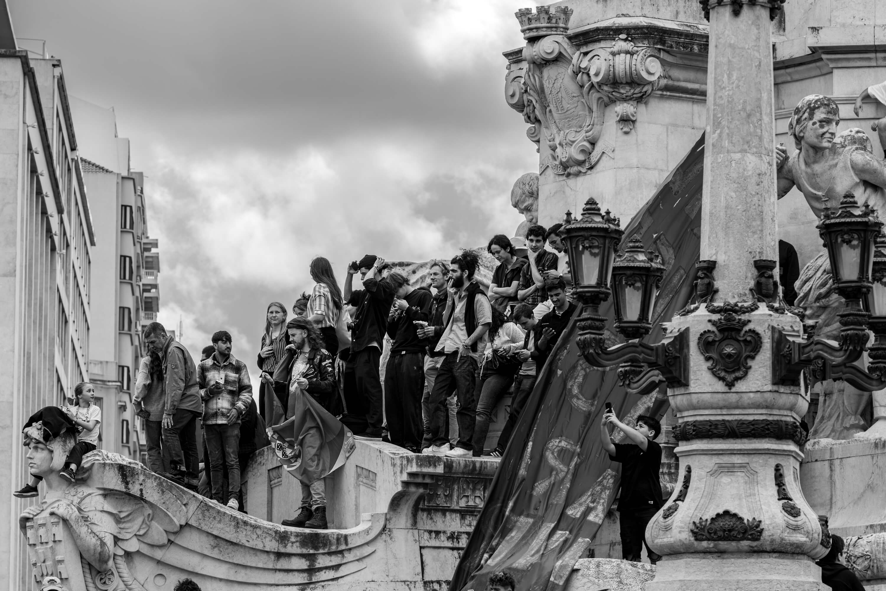 Attendees of the 50th anniversary of the Carnation Revolution climb on top of the statue at Praça Marquês de Pombal. They are all crowding together to get a view of the crowds down below.