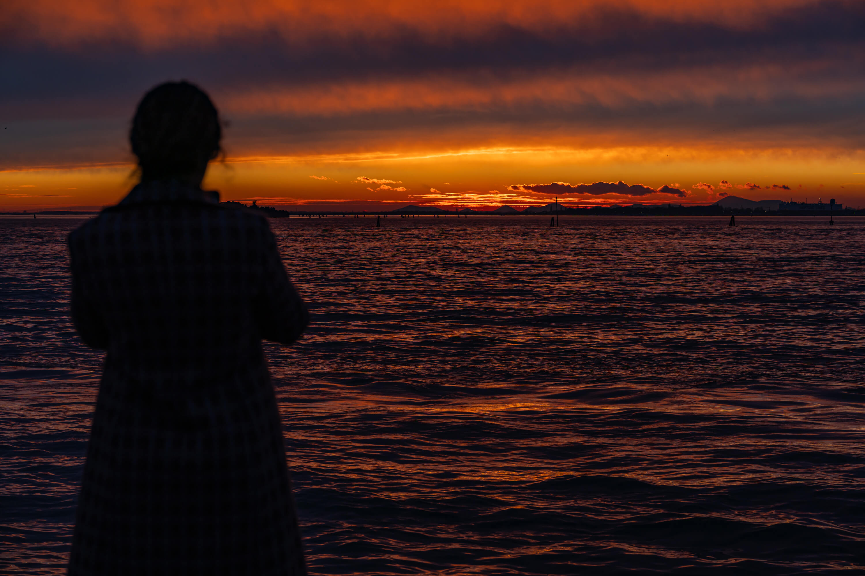 A woman looks out at the final golden light of a sunset. The sky is covered in clouds allowing the light to bounce off them and light up the water, giving the whole scene a look of being on fire.